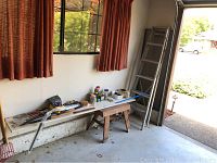 Overview of the workbench along wall with tools laid out on bench and stool beneath; aluminum ladder leaning near corner.