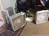 Shows the white oil-filled radiator heater to the left, two white box fans centered and right. Items appear to be well used and placed on carpet and floor near doorway.