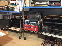 Metal shelving unit with red crate of DVDs on left, black CD carousel on right, and cardboard box of CDs on top shelf