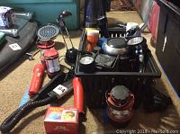 Photo showing assortment of flashlights, lanterns, and a headlamp in a black plastic bin along with items placed beside it on a carpeted floor.