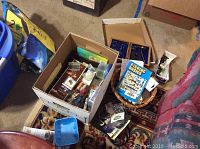 Box filled with assorted fly fishing hooks, lures, and other fishing materials including a fishing basics book and cardboard packaging, placed on a carpet.