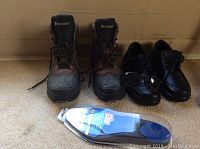 Pair of Mens Columbia hiking boots, black and brown, with laces tied, and next to them black dress shoes along with a pair of blue insoles visible in front of them.