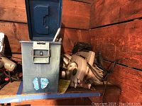 Photo showing several handheld circular saws and saw blades stored in a blue metal box on a blue cart against a wood-paneled wall.