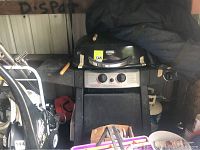 Full view of the black propane BBQ grill with side shelves, showing the two control knobs and partially covered with dark blue cover. Various other outdoor items and a white propane tank are visible.