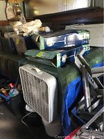 Two new in box Wintair pedestal fans stacked on a covered surface and two white box fans, one standing upright and the other partially obscured.