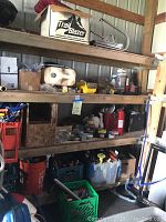 Four-tier wooden shelving unit with various tools and household supplies including plastic crates with hand tools, a small grill on the top shelf, fire extinguisher, and buckets below.