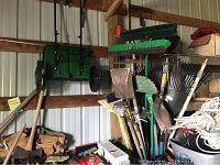 Wide shot showing assortment of yard tools including shovels, rakes, brooms, and rope stored on a shed wall and floor.