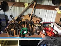 Photo showing variety of hammers, axes, tool belts, tie-down straps, and other hand tools arranged on a table and leaning on a wall.
