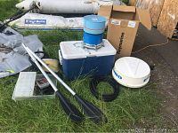 Wide angle shot showing blue cooler, white radar dome, aluminum oars with black blades, tackle box and other marine equipment on grass.