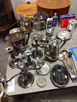 View of multiple silver plated items including coffee pot, candlesticks, serving spoons, bowls, and silver polish bottles on a grey surface.
