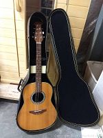 Full view of Ovation 6-string acoustic guitar in its hard shell case with case open, showing wooden top and body shape, resting vertically.