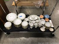 Overview of Noritake Spring Meadow china and accompanying items on black cloth on a cart showing plates, cups, saucers, serving dishes, and iridescent tea set pieces.