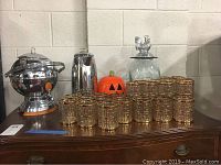 Photo showing entire set on a wooden table, including the large spherical percolator, tall stainless percolator, glass jar with pumpkin lid, and 24 glasses and tumblers with gold geometric patterns.