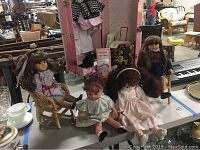 Two Pleasant Company dolls sitting on woven chairs with a smaller third doll, and a doll clothing storage trunk behind them.
