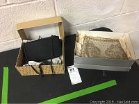 Two clutch purses in original boxes on black surface against concrete wall. Left purse is black with chain strap; right purse is gold-beaded vintage style.