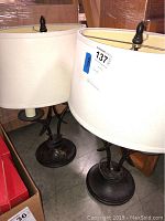 Pair of two-arm metal base lamps with white drum lamp shades shown together from a side angle.