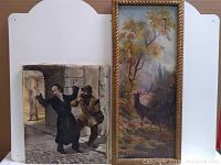 Full view of two paintings leaning against a white board, one depicting two men and the other a stag in forest.