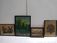 Four framed artworks displayed on a white surface against a white wall, showing two cloth art pieces (with birds and tree designs) and two small paintings of nature and landscapes.