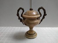 Front view of a marble or onyx urn with two ornate metal handles and a lid, showing the polished stone surface and decorative metal accents.