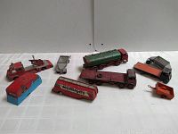 Photo showing 8 vintage Dinky toy vehicles arranged on a table, including various trucks, trailers, and a toy bus with some wear and paint loss visible.