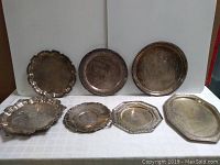 Photo showing all seven silverplate trays arranged side by side on white cloth background. Trays vary in shape and sizes with visible tarnish.