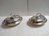 Pair of two silverplate covered dishes with lids and ring handles, one oval and one round, against a plain white background on a grey surface.