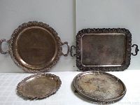 Four vintage silverplate trays arranged standing and lying on a white surface showing size comparison and decorative edges.