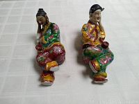 Front view of two porcelain figures sitting with heads resting on hands, displaying colorful painted floral clothing