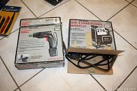 Photo showing boxed cordless drill and manual scroll saw with power cord in box on tiled floor.