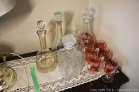 Photo shows three clear glass decanters with stoppers and six red floral wine glasses set on a lace runner on dark wood surface, with two orange vases visible in the background.