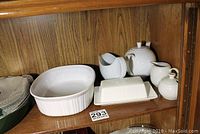White ceramic bakeware and serveware including casserole dish, butter dish, teapot, creamer, sugar bowl, and gravy boat arranged on wooden shelf.