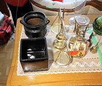 Black cast iron cauldron, black ceramic square vase, brass hanging balancing scales, clear glass vase, green textured glass container, brass candlestick with candle, brass bells arranged on lace runner on wood table