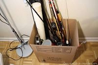 Box filled with assorted fishing rods and reels, various handle materials visible, positioned next to a floor lamp.