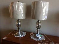 Two 23.5-inch table lamps with metal and glass bases and beige fabric shades, plastic wrapped, sitting on wooden surface with lace doilies.