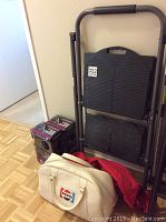Folded black Cosco stepladder with padded handle, four boxed AutoTrends universal bucket seat covers stacked beside it, and three various carrying bags including a white Pepsi bag and red and black bags.