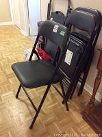 Four black folding chairs standing and stacked against a wall showing front and side views.