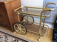 Side view of gold-tone metal bar cart with two large spoked wheels and two glass shelves, showing the cart placed on a wood floor next to furniture.