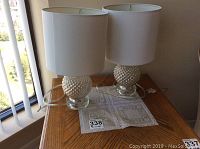 Pair of two similar glass base lamps with white shades on a wooden side table by a window.