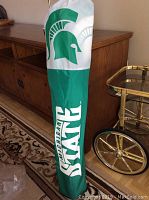 Photo of the green and white carrying case for the Michigan State folding chair with Spartan helmet logo and 'Michigan State' text.