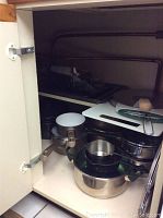 Various stainless steel pots and pans stacked inside kitchen cabinet, including one with white non-stick interior and several transparent lids