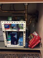 Photo shows kitchen storage under sink with white shelf displaying boxes of Ziplock storage and freezer bags, plastic wrap, stove burner covers, and a box of Finish dishwasher cleaning cubes.