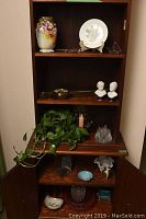 Full view of five shelf wooden shelving unit with various decorative items arranged on all shelves, including vase, plate, busts, plant and ceramics.