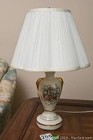 Full view of lamp showing white pleated shade atop ceramic base with decorative classical scene and gold accents.