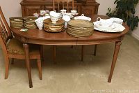 Full view of the oval oak dining table with various porcelain dishware placed on top and two upholstered dining chairs visible.