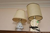 Two vintage lamps with white ceramic bases and beige fabric shades placed side by side on a shelf with a live Schefflera plant.