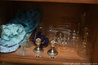 Image showing six silver-plate items including two candlesticks and holding bags in a cabinet with vintage crystal stem wine glasses behind them.