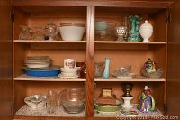 Two wooden kitchen cupboards filled with assorted glassware, plates, bowls, a CorningWare skillet, and decorative pottery items.
