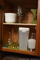 View of cabinet shelf showing white ceramic bowl, clear glass vintage coffee pot, green plasticf floral frog, white plastic container with lid, white soap dispenser with metal base, and an assortment of glass bud vases.