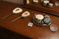 Photo showing hand mirror, hairbrush, glass tray with cosmetic items, and small glass bowl arranged on wooden dresser in front of large mirror.
