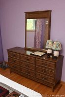 Full view of the brown wood veneer dresser with nine drawers and matching wood-framed mirror against purple wall.